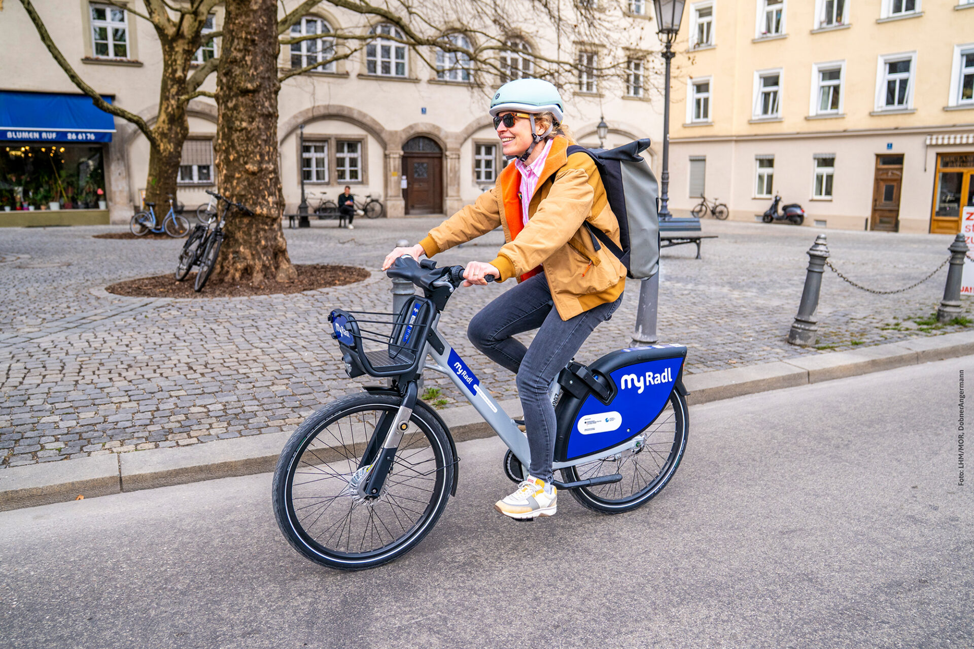 Eine Frau mit Radhelm und Rucksack fährt lächeln auf ihrem MyRadl Leihrad eine Straße entlang.